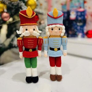Puede incluir: Dos muñecos cascanueces hechos a mano con ganchillo. Uno es rojo, verde y dorado, el otro es azul, rojo y dorado. Ambos tienen pelo y bigote blancos, cinturones negros y llevan sombreros. Están de pie frente a un árbol de Navidad.