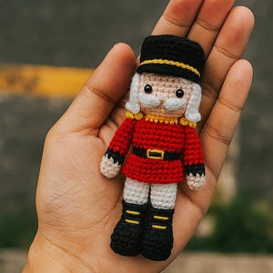 May include: A small, handmade crochet nutcracker doll. The doll wears a red jacket with gold trim, a black hat with a yellow band, and black boots. It has a white mustache and hair. The doll is approximately 10 cm tall.