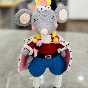 Op de afbeelding: Een grijze gehaakte muis die een roze, rood en geel koninklijk kostuum draagt met een kroon. De muis heeft een roze neus en roze oren met witte accenten.