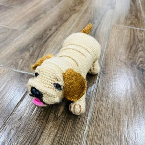 Puede incluir: Un juguete de perro tejido a ganchillo en crema y marrón. El perro tiene orejas marrones, una nariz negra y una lengua rosa. El juguete está sobre una superficie de madera.