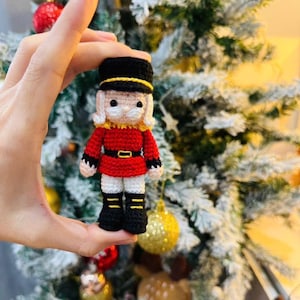 Puede incluir: Muñeca cascanueces de ganchillo hecha a mano. La muñeca lleva una chaqueta roja, botas negras y un sombrero negro con una banda dorada. La muñeca tiene una cara blanca y un cuello amarillo. El fondo es un árbol de Navidad.