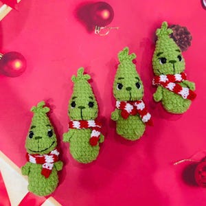 May include: Four green crocheted Grinch ornaments with red and white striped scarves. The ornaments are on a pink background.
