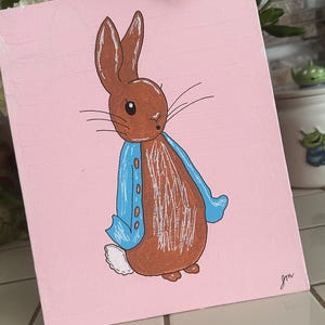 May include: A hand-painted artwork on a pink background features a brown rabbit wearing a blue jacket. The rabbit has long ears and whiskers. The artwork is signed in the bottom right corner. The painting is displayed on a white surface.