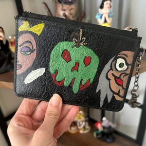 May include: A black leather zippered pouch with a hand-painted design featuring the Evil Queen, Snow White's poisoned apple, and the Evil Queen's reflection. The pouch has a silver chain attached.