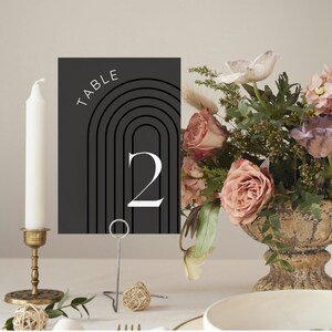 Op de afbeelding: Zwarte tafelnummerkaart met witte tekst die "TABLE" en "2" aangeeft met een geometrisch ontwerp in zwart en wit. De kaart staat op een metalen standaard en voor een bloemstuk.