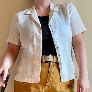 May include: A cream-colored short-sleeved button-down shirt paired with mustard-yellow cargo pants. The outfit includes a black top and a light-colored belt. The pants have large pockets on the sides.