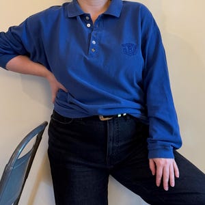 May include: A long-sleeve blue polo shirt featuring a collar, a button-up placket with four visible buttons, and a dark embroidered crest logo on the left chest area. The shirt is paired with black denim jeans featuring a black belt with a silver buckle.