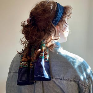 May include: A dark blue headscarf with white polka dots and a forest print scarf tied in the hair. The scarf is paired with a denim shirt. The hair is curly and brown.