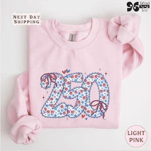 May include: Light pink sweatshirt with a floral "250" design in red and blue. The design includes bows and hearts. The text "Next Day Shipping" is at the top left, and "Light Pink" is at the bottom right.