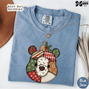 Peut inclure: Un t-shirt bleu clair avec un motif de décoration de Noël représentant un visage d'ours en patchwork. L'ornement a un capuchon doré et des feuilles de houx. Un collier doré est drapé sur le t-shirt. Le t-shirt porte l'inscription "Blue Jean".