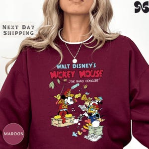 A person wearing a maroon sweater with a graphic design featuring Mickey Mouse and other Disney characters from Walt Disney's "Mickey Mouse" concert.