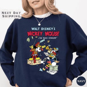 A navy blue sweatshirt with a graphic design featuring Mickey Mouse and other Disney characters, along with the text "Walt Disney's Mickey Mouse" and "The Band Concert".