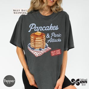 May include: Dark grey t-shirt featuring the text "Pancakes & Panic Attacks" above a pancake illustration with butter and syrup, served with blueberries on a plate. A red and white checkered napkin is beneath the plate. The shirt has a "Buy One Get One" sticker.