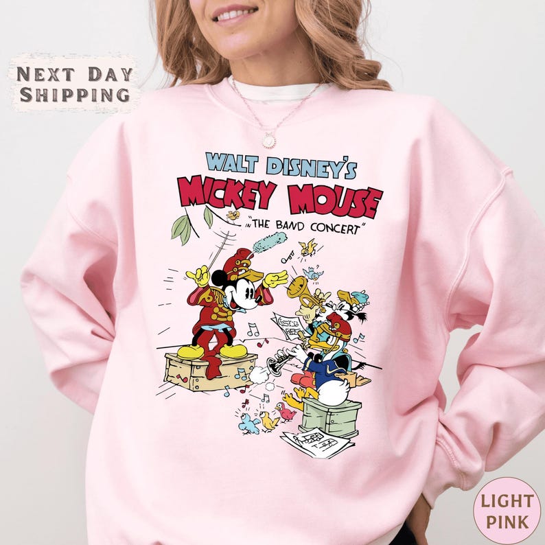 A woman wearing a pink sweatshirt with a Disney Mickey Mouse graphic and text.
