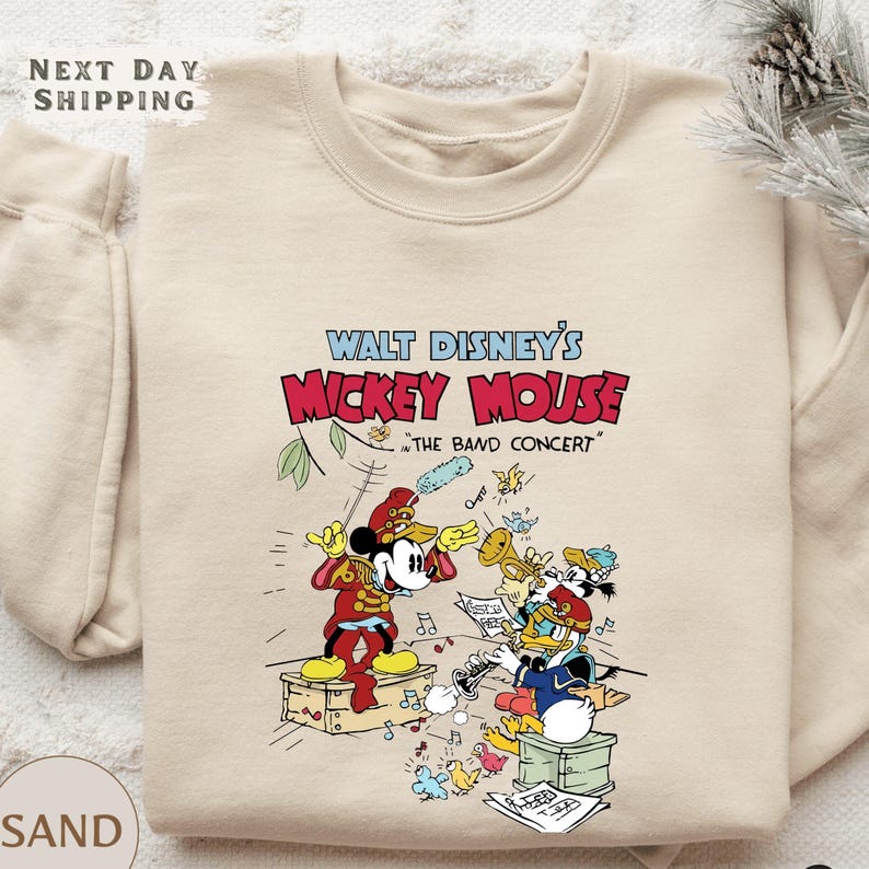 A beige sweatshirt with a colorful graphic design featuring Mickey Mouse and other Disney characters, along with text that says "Walt Disney's Mickey Mouse 'The Band Concert'".