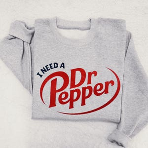 May include: A light gray sweatshirt with the text "I NEED A Dr Pepper" in red and blue. The Dr Pepper logo is prominently displayed in a red, stylized font.