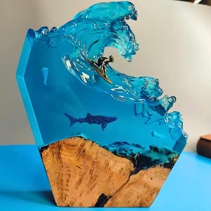 Puede incluir: Una escultura de resina azul de una ola rompiendo sobre un surfista y un tiburón ballena. La base de la escultura es de madera.