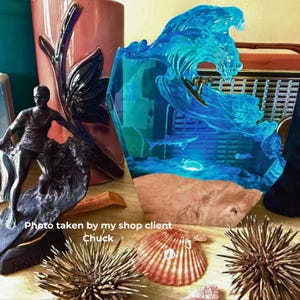 Puede incluir: Una escultura de resina azul de una ola con un surfista. La ola es translúcida, revelando un paisaje urbano y vida marina. También se ve una estatua de surfista de bronce y otra decoración con temática oceánica.