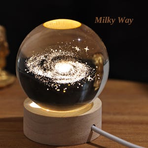 Pode incluir: Um globo de vidro com um design de galáxia em espiral no seu interior, iluminado por baixo por uma base de madeira. O globo está etiquetado como "Via Láctea".