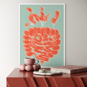 May include: An abstract art print with a coral-colored design on a light teal background, framed in white. A pink teacup, a decorative sphere, a small white tray with candles, and a book are on a red-brown cabinet.