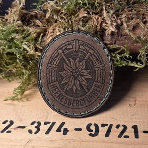 May include: A circular brown leather patch with an embossed design of a flower and the text "HEERESBERGFUHRER". The patch has a green stitched border. The background includes moss and a wooden surface with printed numbers.