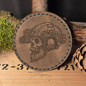 Könnte beinhalten: Ein runder, brauner Lederaufnäher mit einem detaillierten gravierten Bild eines Schädels, der einen Militärhelm mit Nachtsichtgeräten trägt. Der Aufnäher hat einen blau genähten Rand und ist vor einem natürlichen Holzhintergrund platziert.