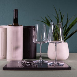 Könnte beinhalten: Zwei weiße Weinkühler mit einer Weinflasche in der Mitte, zwei klare Weingläser, ein schwarzer Weinkühler und ein Korkenzieher auf einer schwarzen Oberfläche.