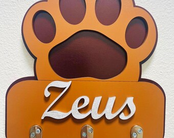 May include: A wooden wall-mounted key holder in the shape of a dog paw. The paw is orange with a dark brown interior. Below the paw is a rectangular section with the name "Zeus" in silver script, and three silver hooks.