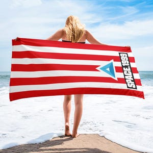 May include: A red and white striped beach towel with the text "SPACE.1999" in white. The towel features a blue triangle design. The towel is being held up by a person on a beach.