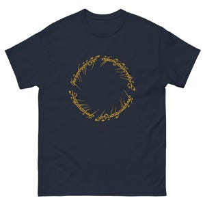 May include: Navy blue t-shirt featuring a gold-colored circular design with stylized text. The short-sleeved shirt is made of a soft material. The design is centered on the front of the shirt.
