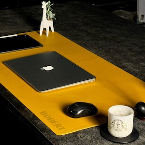 Puede incluir: Una alfombrilla de escritorio de color amarillo mostaza con el nombre "ROBERT" impreso. Una computadora portátil, un ratón y una tableta negros están sobre la alfombrilla. Un macetero blanco con forma de jirafa, una vela, una pipa y una planta pequeña también están en el escritorio.