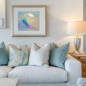 Puede incluir: Un sof&aacute; blanco con almohadas de estampado azul y blanco. Un cuadro enmarcado de una ola con una puesta de sol en el fondo cuelga de la pared sobre el sof&aacute;. Una l&aacute;mpara con una pantalla blanca se encuentra en una mesa junto al sof&aacute;.