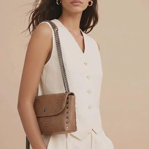 Peut inclure: Sac bandoulière marron texturé avec une chaîne et des clous décoratifs. Le sac a un rabat et une pampille. Il est porté par une personne vêtue d'un gilet et d'un pantalon blancs.