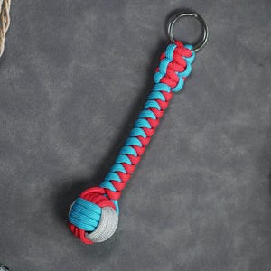 May include: A red and blue braided rope keychain with a knotted ball at the end. The keychain is attached to a silver key ring.