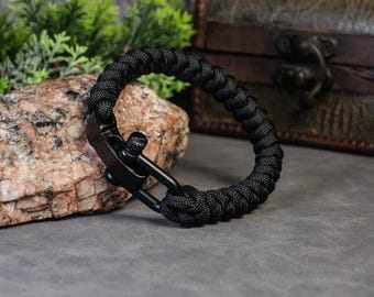 May include: Black paracord bracelet with a metal clasp. The bracelet is made of tightly woven black cord and features a secure, matte black clasp. The bracelet is shown on a rock surface.