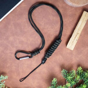 May include: Black and green paracord lanyard with a metal carabiner clip. The lanyard is made of woven cord and features a loop at the end. The carabiner is silver in color.