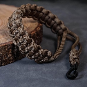 May include: A brown paracord camera strap with a black metal hook. The strap is made of braided cord and has a simple design.