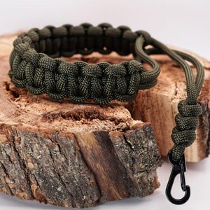 May include: A dark green paracord bracelet with a black metal clip. The bracelet is made with a braided paracord and has a simple design.