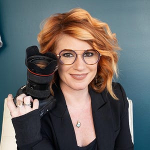 A woman with red hair and glasses is smiling and holding a camera in front of her face.