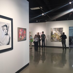 an art gallery with several people standing and observing the artworks on display. The artworks include framed pictures and paintings on the walls, creating an atmosphere of appreciation and engagement.