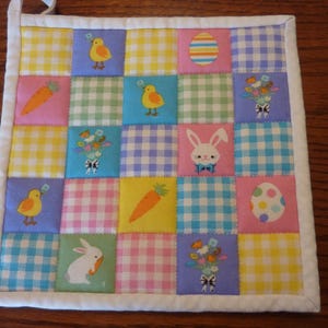 May include: A patchwork Easter-themed hot pad with a white border and hanging loop. The squares feature gingham patterns and images of chicks, carrots, bunnies, flowers, and Easter eggs. The color palette includes yellow, pink, blue, and green.