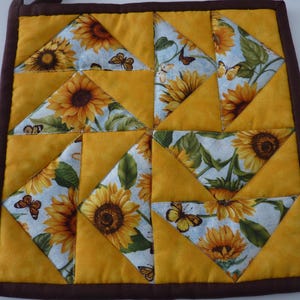 May include: A square quilted potholder featuring a patchwork design of solid mustard yellow triangles and a white fabric print with yellow sunflowers, green leaves, and brown butterflies, all bordered by a dark brown fabric binding.