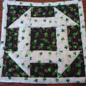 May include: A white fabric pot holder with a black and green shamrock pattern. The pot holder is square and has a stitched border.