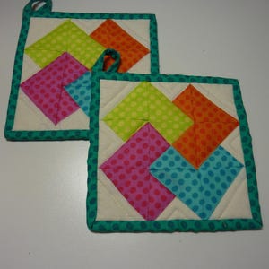 May include: Two square quilted pot holders with colorful geometric designs. Each features a cream-colored background with a pattern of bright pink, blue, orange, and green polka-dotted squares. The edges are trimmed with a teal fabric.