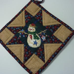 May include: A square quilted potholder featuring a central dark green square with a snowman illustration. The design includes a star pattern made of tan burlap-textured fabric and a dark blue fabric with small red hearts and blue stars.