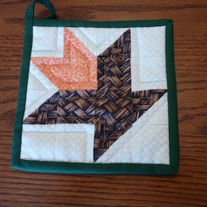 May include: A green-trimmed pot holder with a brown and orange patchwork design. The design features a leaf shape with a woven pattern.