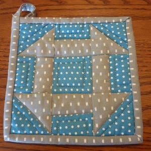 May include: Square quilted potholder with a loop for hanging. The design features a patchwork pattern of gray fabric with white polka dots and teal blue fabric with white polka dots. The edges are gray with white dots.