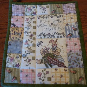 May include: A green quilted hot pad with a patchwork design. The center square features a floral design with the text "Plant smiles grow giggles harvest love".