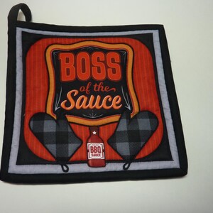 May include: Square, quilted pot holder with a black border and a loop for hanging. The center features the text "BOSS of the Sauce" in orange and black, with a red background. Two oven mitts and a BBQ sauce bottle are also shown.
