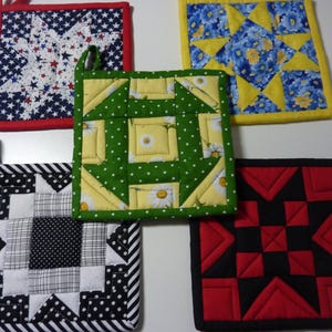 May include: Five square quilted pot holders with various patterns. One has a red, white, and blue star design. Another features a green background with yellow and white floral accents. The others have black and white, red and black, and blue and yellow designs.